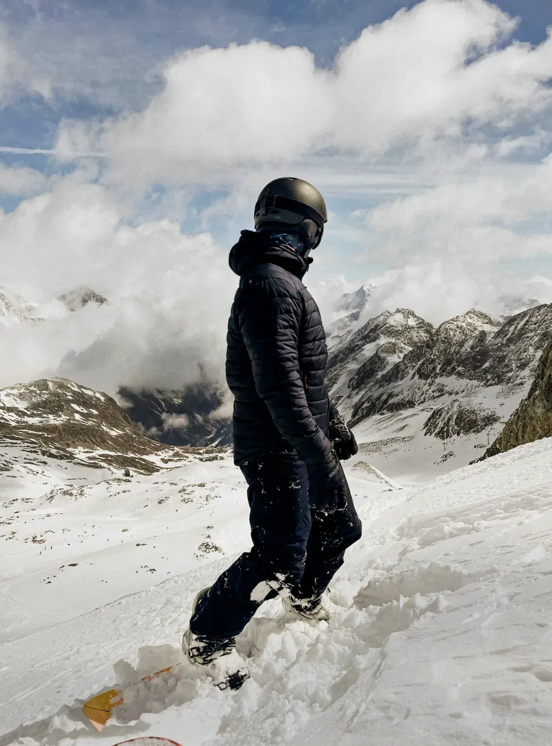 A snowboarder in a black insulated jacket on a snowy ridge above the clouds
