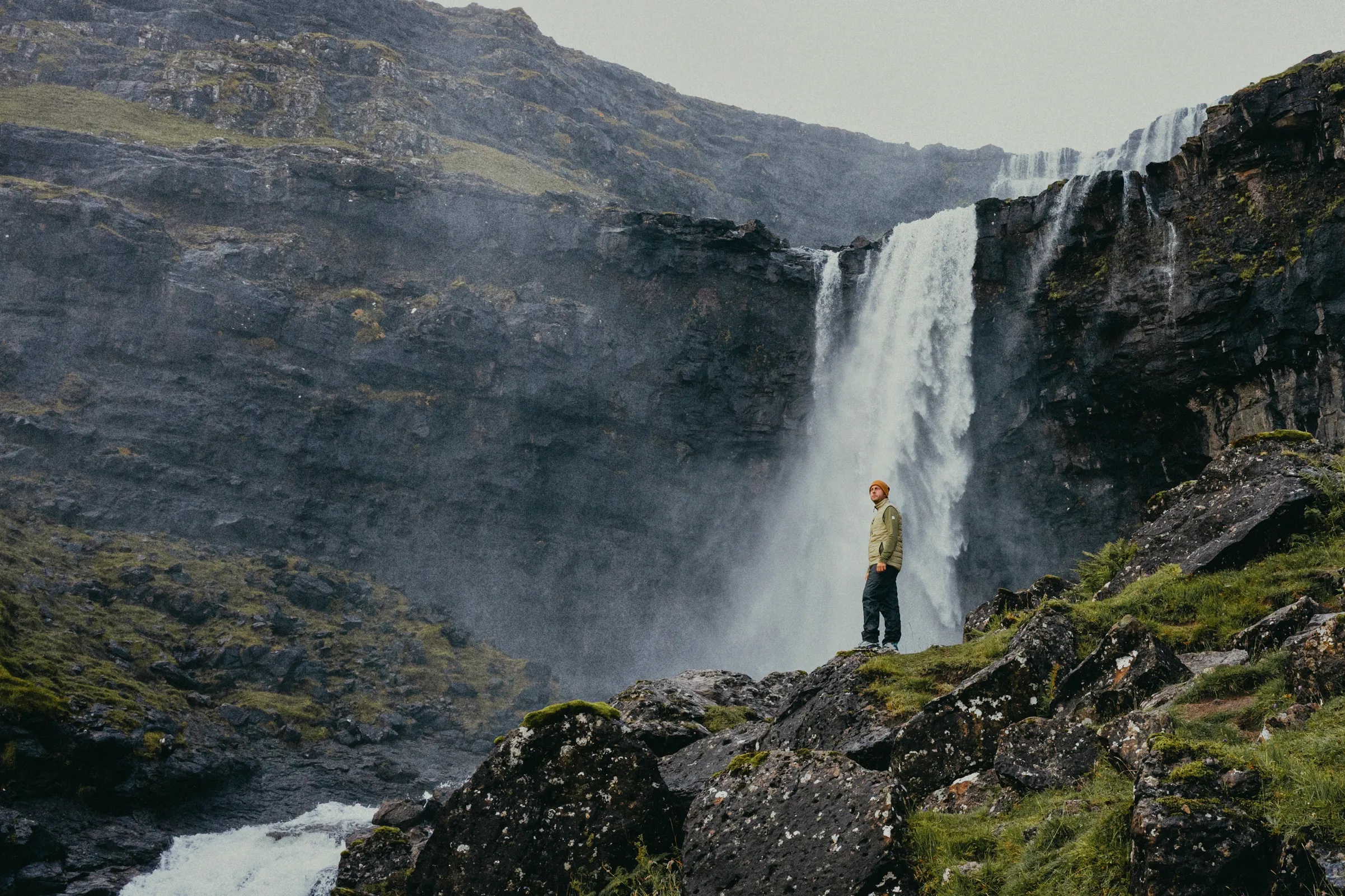 A hiker in a quilted vest and orange beanie standing on mossy rocks below a tall waterfall in a dark cliff face