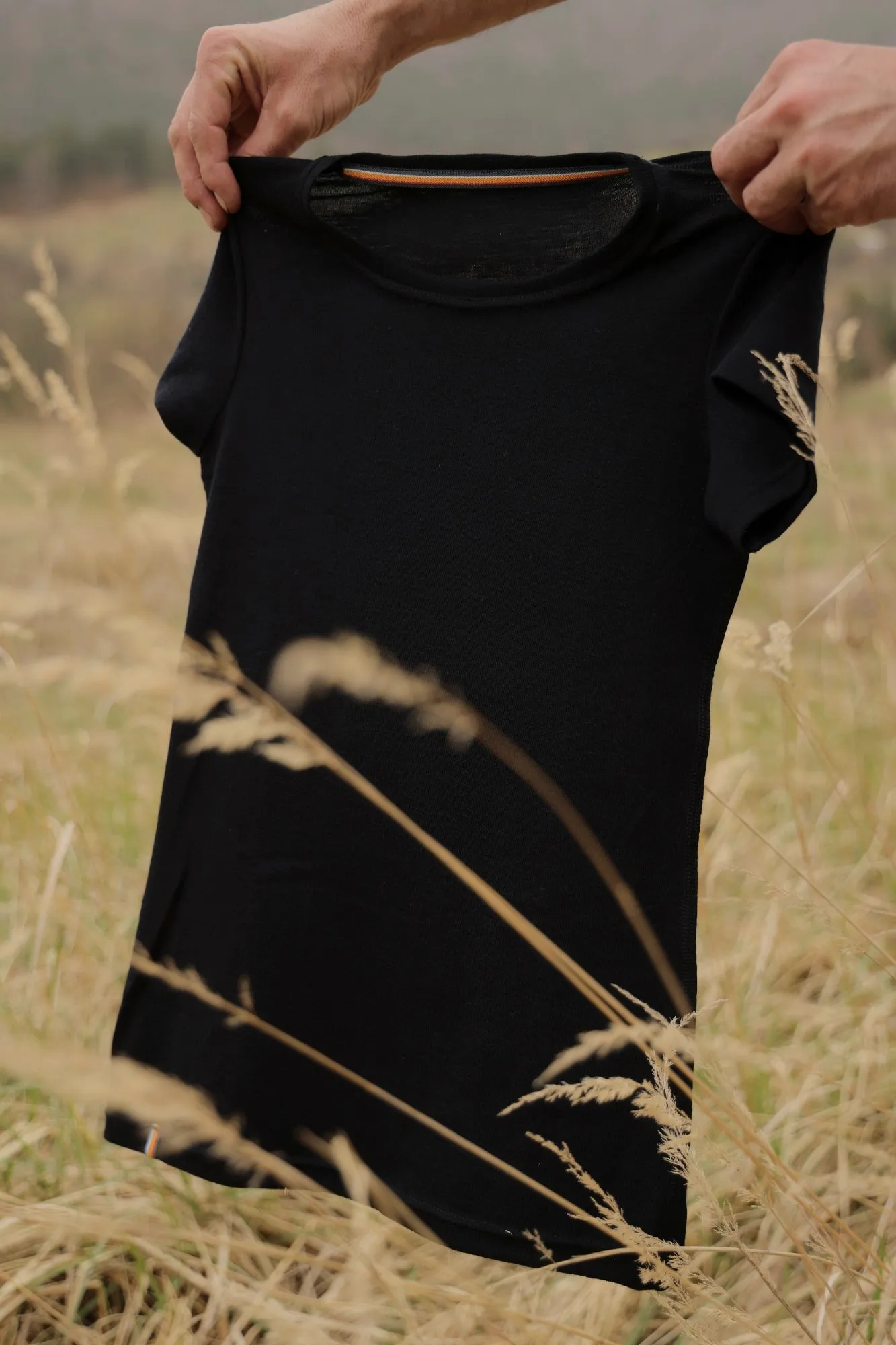 Two hands holding up a black merino T-shirt in a field of tall dry grass