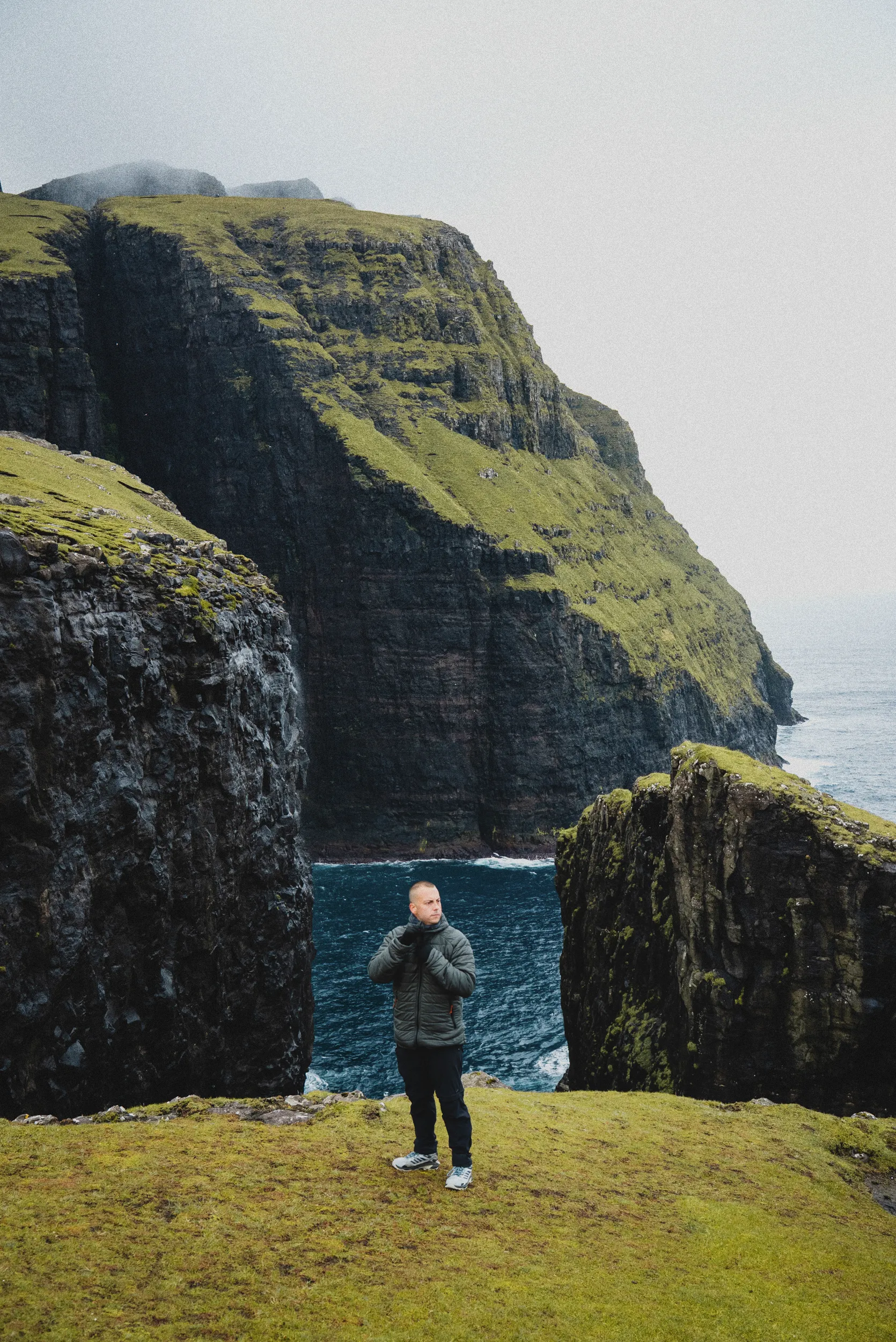A man in a grey puffer jacket standing on a grassy clifftop in front of a narrow sea inlet between dark, moss-covered cliffs